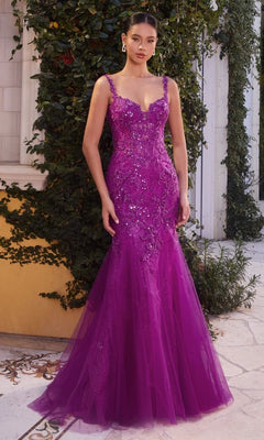 A woman stands outdoors in the Long Lace Mermaid Prom Dress: Andrea and Leo A1231, a fitted, floor-length purple gown with floral appliqué, sequin details, and a sheer bodice. Green vines cover the wall behind her.