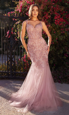 A woman stands outdoors in front of a gate and flowering bushes, wearing the Andrea and Leo A1231 Long Lace Mermaid Prom Dress in light pink with embroidered lace accents and a sheer tulle skirt.