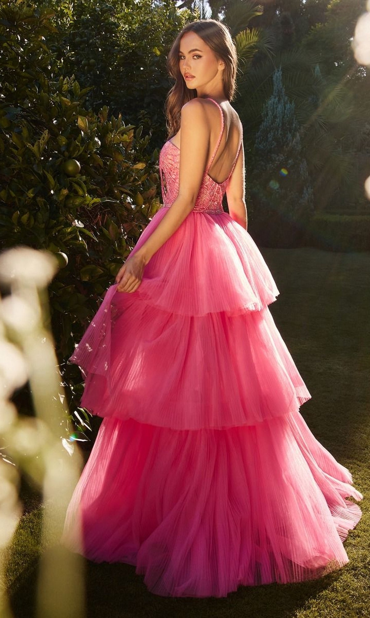 A woman stands outdoors in a garden, wearing the High-Low Beaded-Bodice Ruffle Prom Ball Gown A1239 in bright pink with a fitted, sleeveless, open-back bodice, looking over her shoulder.