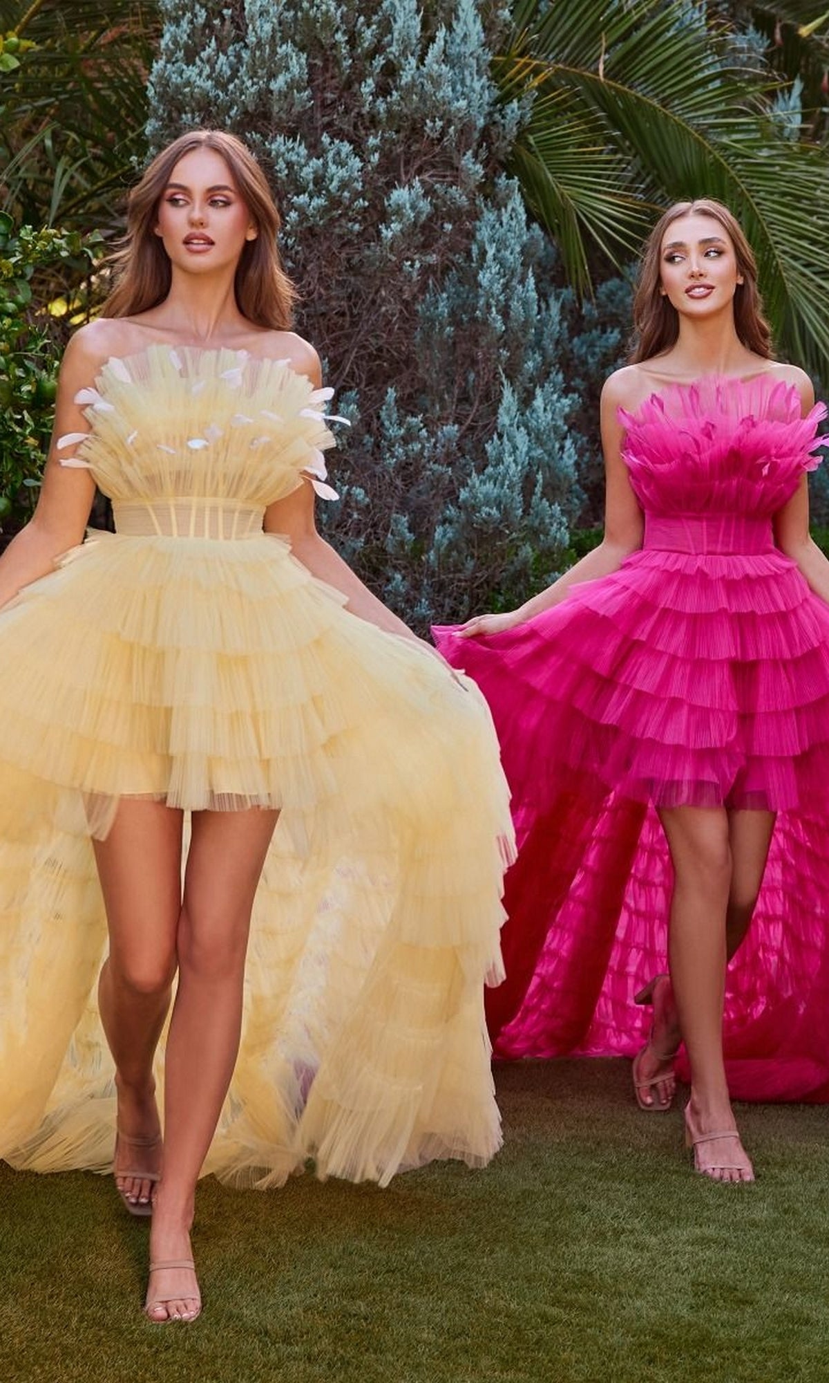 Two women stand outdoors among lush greenery, one in a pale yellow strapless ball gown and the other in the Ruffled High-Low Ball Gown A1240 by Andrea and Leo—a bright pink ruffle a-line dress.