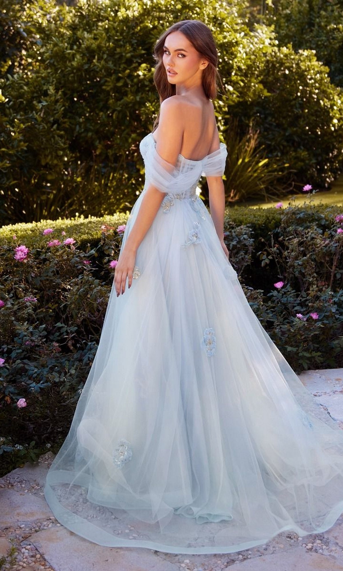 Outdoors in a garden, a woman wears the Andrea and Leo A1246 Long Blue Prom Ball Gown, featuring floral embellishments and a flowing tulle skirt, as she glances back over her shoulder.
