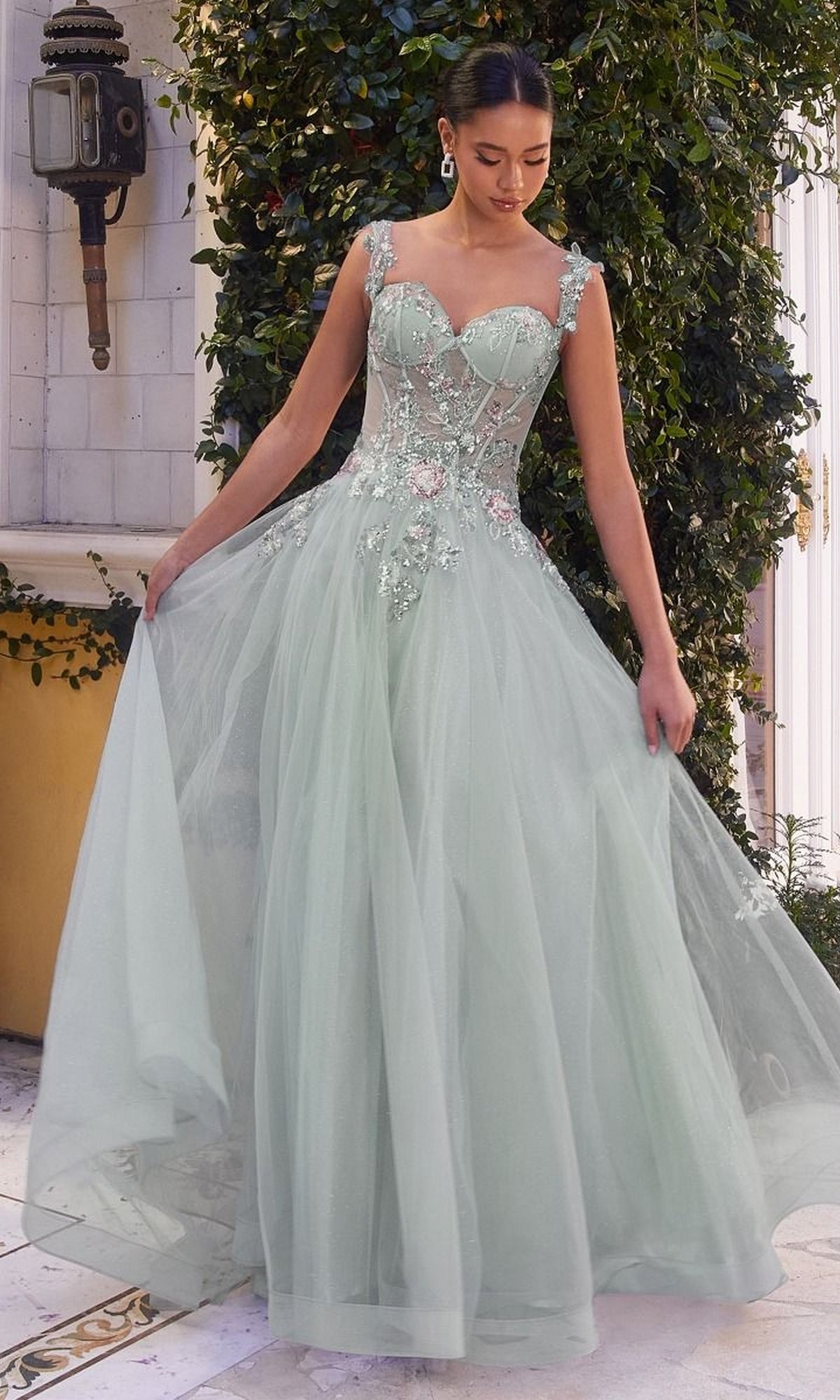 A woman in the Long Pastel A-Line Prom Dress (Andrea & Leo 1258), featuring light green embroidered tulle, stands outdoors by greenery, gracefully holding her skirt.
