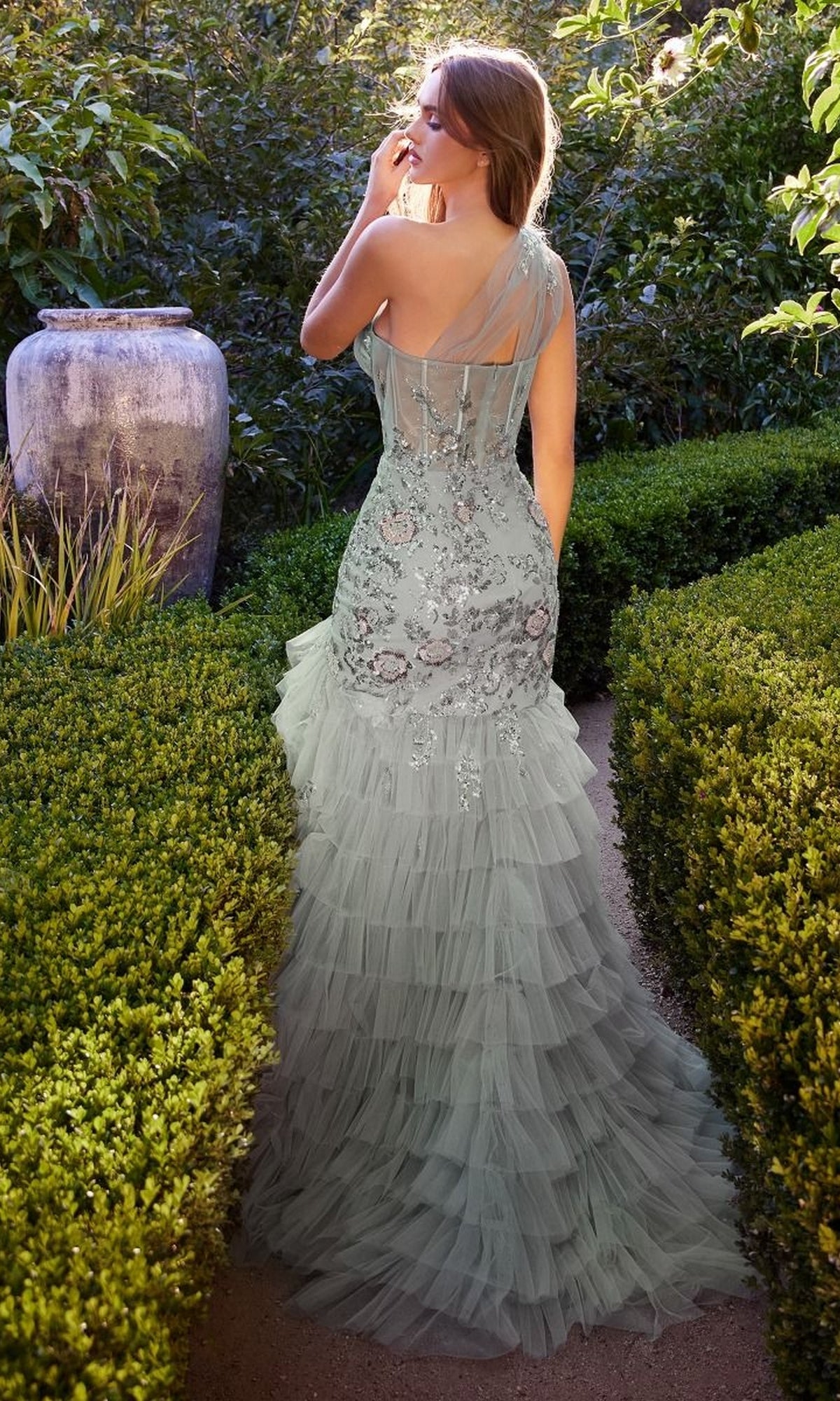 A woman in the Andrea and Leo A1260 long ruffle green prom dress stands in a garden, surrounded by manicured hedges and lush greenery, her back turned to the camera.