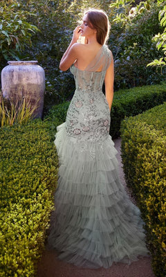 A woman in the Andrea and Leo A1260 long ruffle green prom dress stands in a garden, surrounded by manicured hedges and lush greenery, her back turned to the camera.