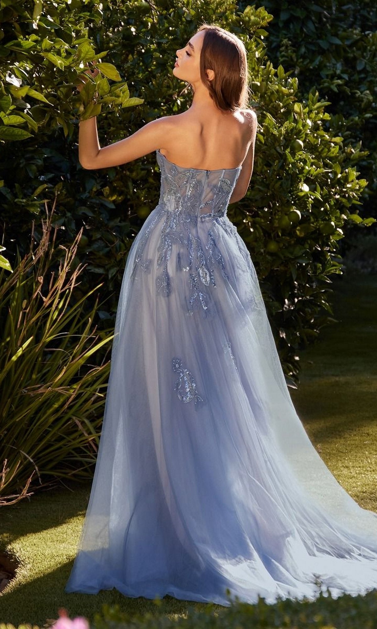 A woman stands in a garden wearing the Andrea & Leo A1294 long blue prom ball gown with floral embellishments, the strapless formal dress flowing gracefully to the ground as she faces away from the camera.