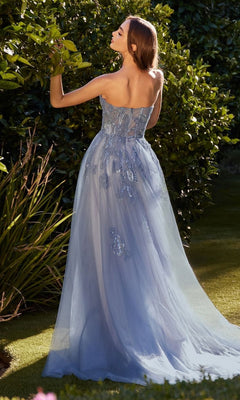 A woman stands in a garden wearing the Andrea & Leo A1294 long blue prom ball gown with floral embellishments, the strapless formal dress flowing gracefully to the ground as she faces away from the camera.