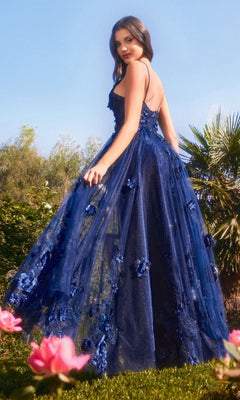 Wearing the Andrea and Leo A1326 Long Glitter Prom Ball Gown, a woman stands among pink flowers and greenery, glancing over her shoulder in a shimmering blue dress with floral embellishments.
