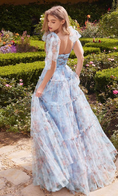 A woman wears the Blue Print Long Prom Dress with Bow Straps A1332, standing on a garden path amid green hedges and blooming flowers.