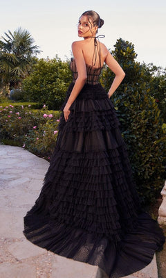 A woman wearing the Long Black Ruffled Prom Gown: Andrea and Leo A1335 stands outdoors on a stone path amidst greenery and blooming flowers, facing away from the camera.