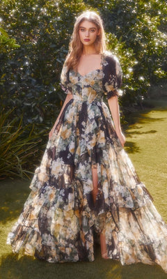 A woman stands on grass, sunlight filtering through greenery, wearing the puff-sleeve long prom dress Andrea and Leo A1336—a formal floral-print gown with ruffled details.