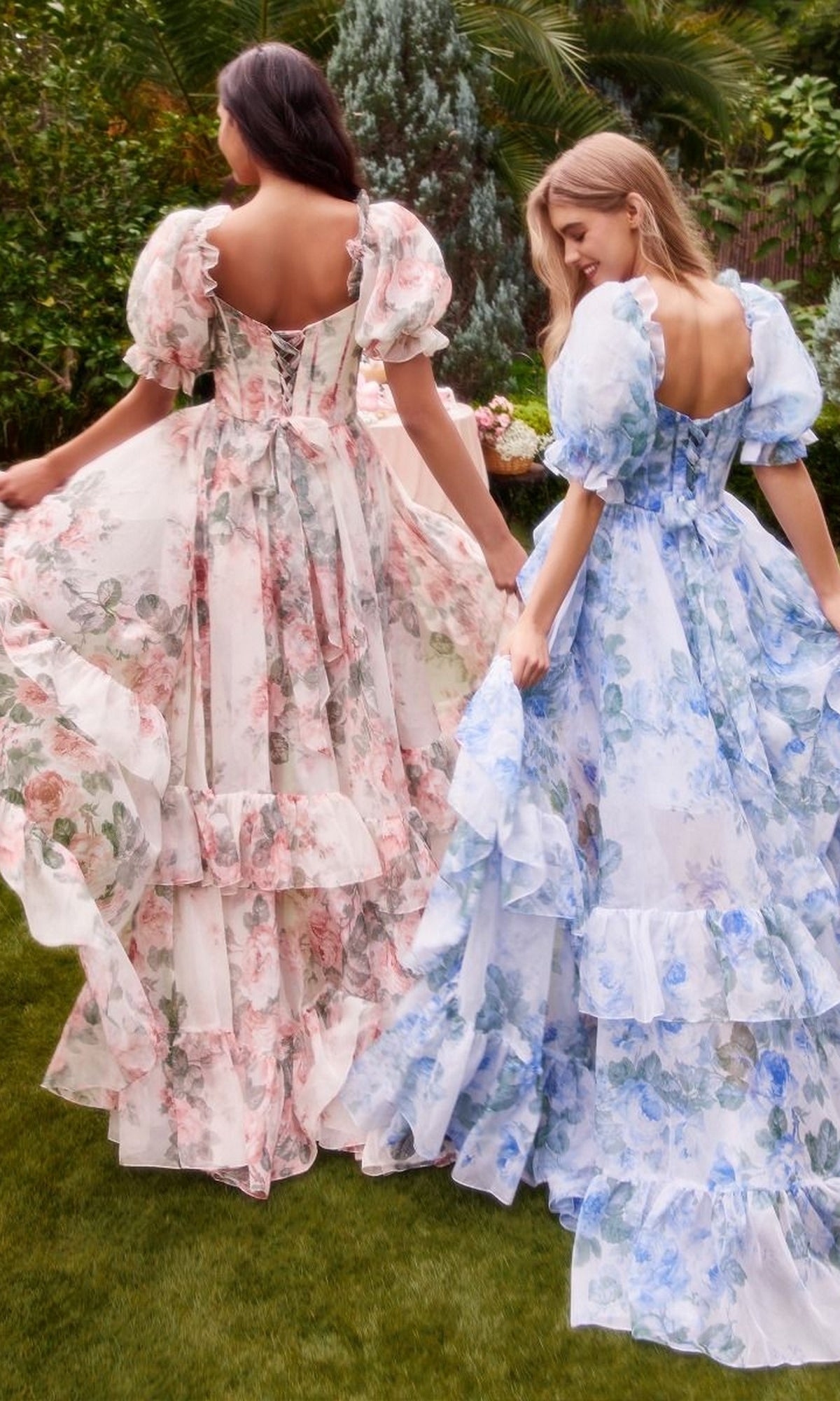 Two women in Andrea and Leo A1336 puff-sleeve long prom dresses stand on grass, holding their skirts, surrounded by lush greenery and trees.