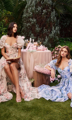 Two women wear the Andrea and Leo A1336 puff-sleeve long prom dresses, sitting in a garden by a table with pink teapots and cups, surrounded by lush greenery and blooming flowers.