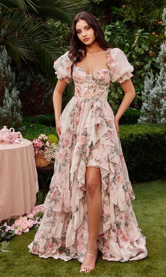 Outdoors, a woman wears the Andrea and Leo A1336 Puff-Sleeve Long Prom Dress—pink, floral, and ruffled—beside a table with a tea set and flowers amid lush greenery.
