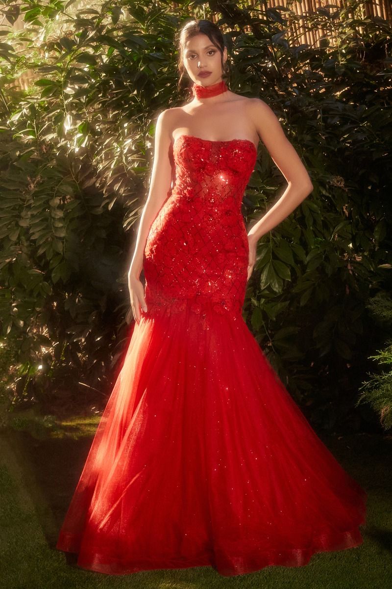 A woman stands outdoors amid lush greenery wearing the Red Prom Dress A1345 by Andrea and Leo, a strapless mermaid gown with a fitted bodice, flowing floor-length skirt, and a matching red choker.
