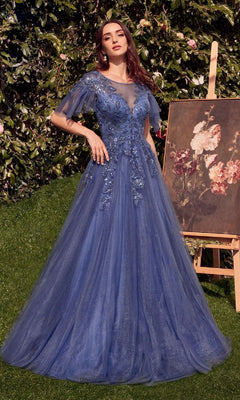 A woman in the Andrea & Leo A1351 Short-Sleeve Prom Ball Gown with blue lace and tulle stands on grass by an easel displaying a floral painting, surrounded by lush greenery.