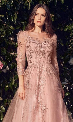 Wearing the Andrea & Leo A1352 Dusty Rose Pink Formal Dress with long sleeves and delicate embroidery, a woman stands before lush green foliage, gazing softly to the side.