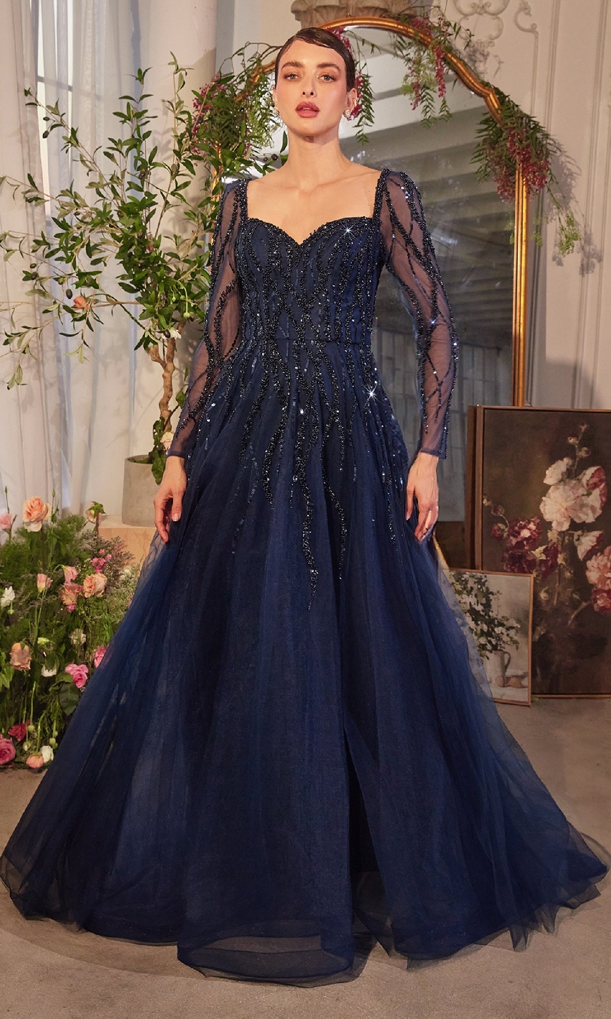 A woman in the Andrea & Leo A1367 Long Sleeve Prom Ball Gown, a navy blue beaded evening dress, stands indoors near floral arrangements and a large gold-framed mirror.
