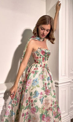 Wearing the Andrea and Leo A1425 long floral-print prom dress, a woman stands indoors by a white paneled wall, looking down with one arm raised. The floor-length gown’s colorful pattern brings elegance to her look.