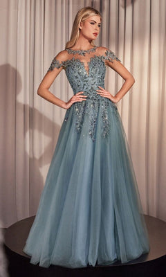 A woman wears the Andrea and Leo A1475 long prom dress in dusty blue, featuring floral appliques on the bodice and sheer cap sleeves, standing before beige curtains.