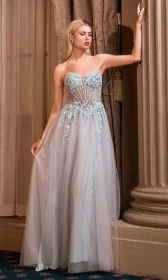 A woman stands indoors in a formal strapless blue beaded prom gown—Andrea & Leo A1478—featuring a sheer skirt and floral-embellished bodice.