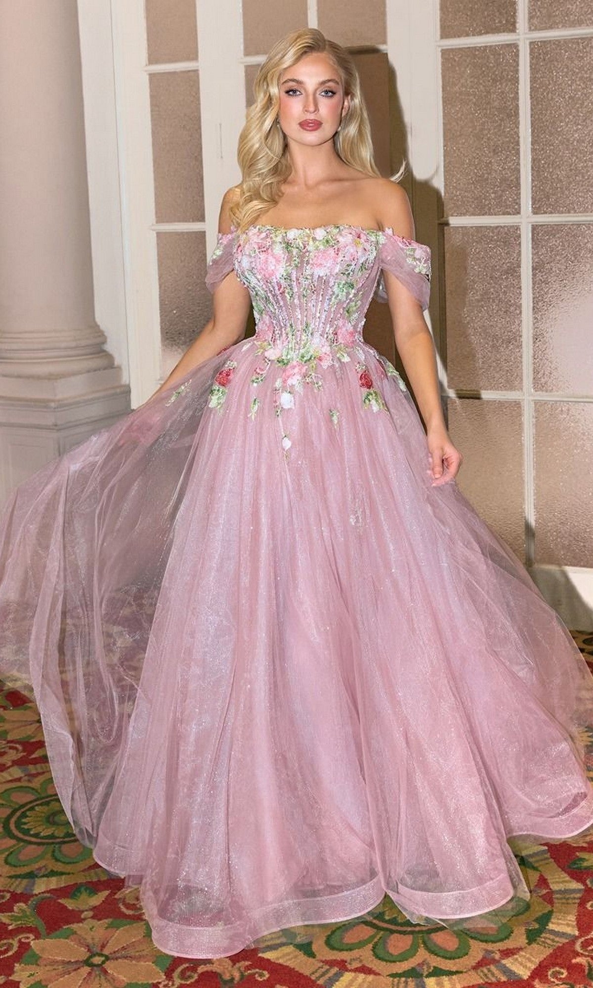 A woman stands indoors on a patterned carpet near glass-paneled doors, wearing the Andrea and Leo A1479 mauve pink prom ball gown.