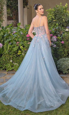 A woman stands in a garden wearing the Long Blue Prom Ball Gown: Andrea and Leo A1490, a light blue embroidered dress with floral details, semi-sheer fabric, and a flowing train, as she looks back over her shoulder.