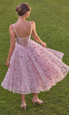 A woman stands on grass wearing the Andrea & Leo A1523 midi prom dress, a light pink floral lace design featuring a sheer corset bodice with a lace-up back and delicate spaghetti straps, paired with matching heels.