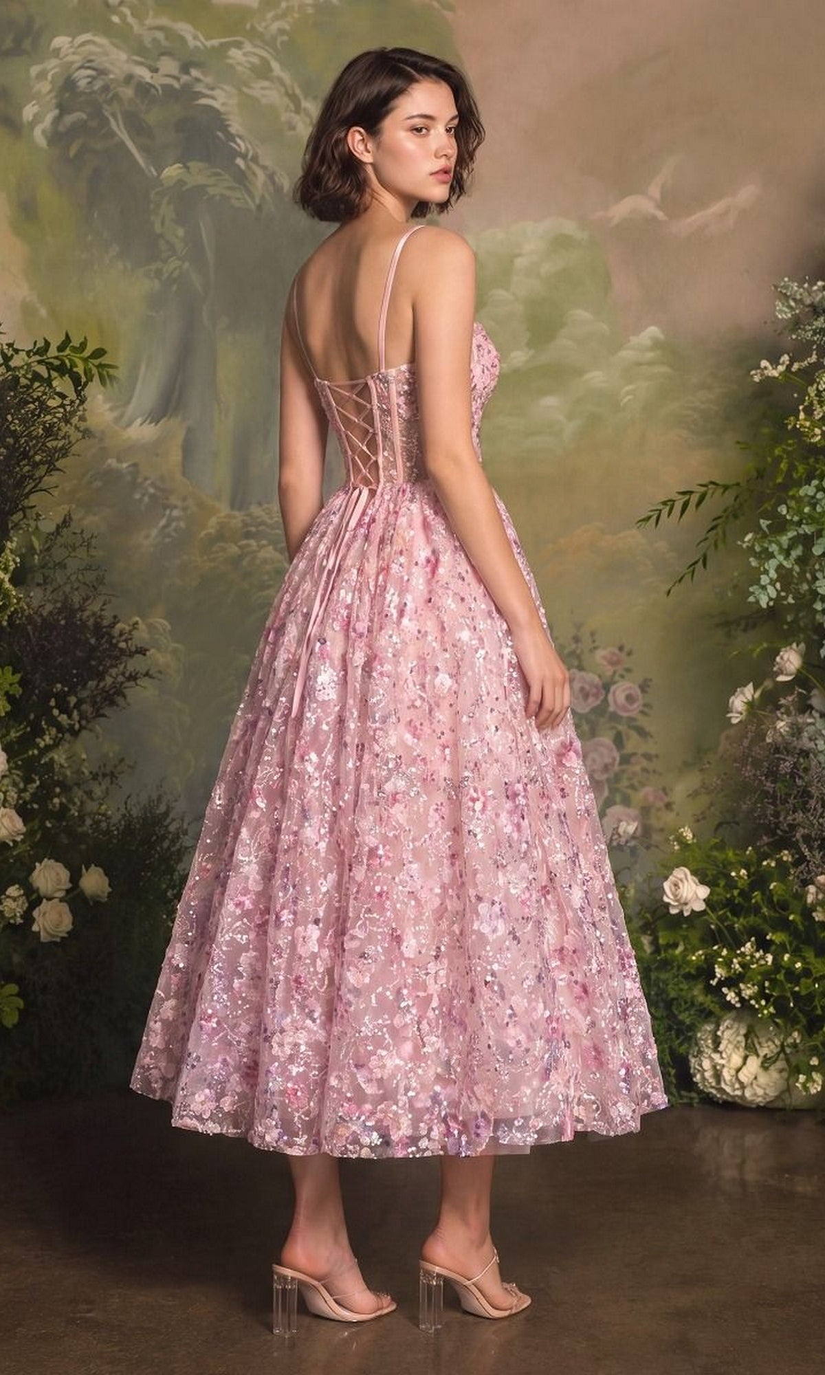 A woman wearing the Andrea & Leo A1523 midi prom dress, featuring pink fabric, sheer panels, and delicate 3D floral appliques, stands in front of a painted garden backdrop.