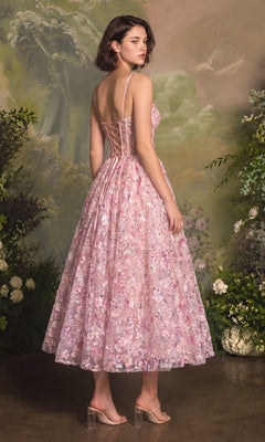 A woman wearing the Andrea & Leo A1523 midi prom dress, featuring pink fabric, sheer panels, and delicate 3D floral appliques, stands in front of a painted garden backdrop.