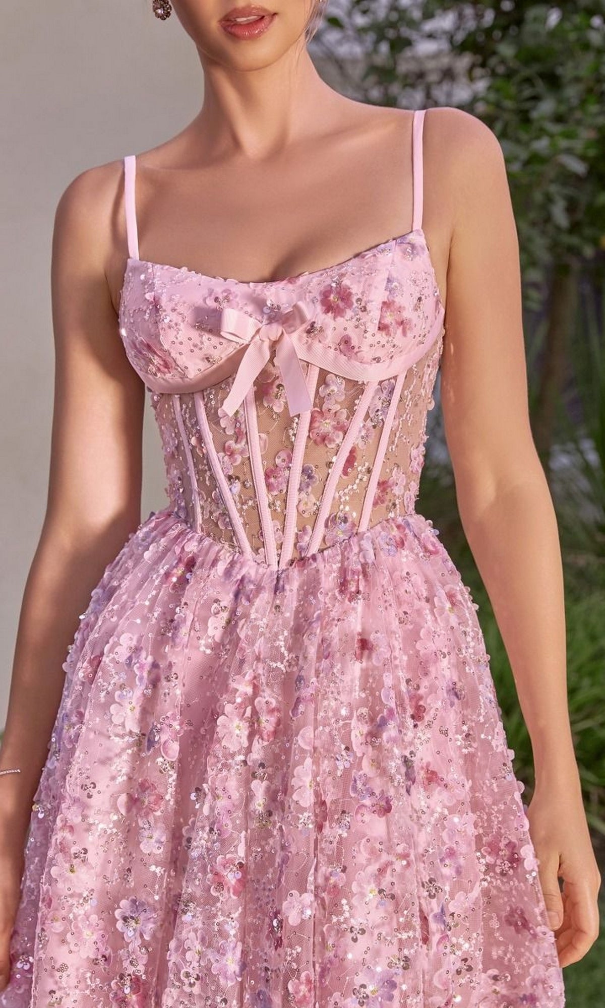 A woman wearing the Andrea & Leo A1523 midi prom dress, a sleeveless pink sequin design with a sheer corset-style bodice, thin straps, and a bow at the bust.