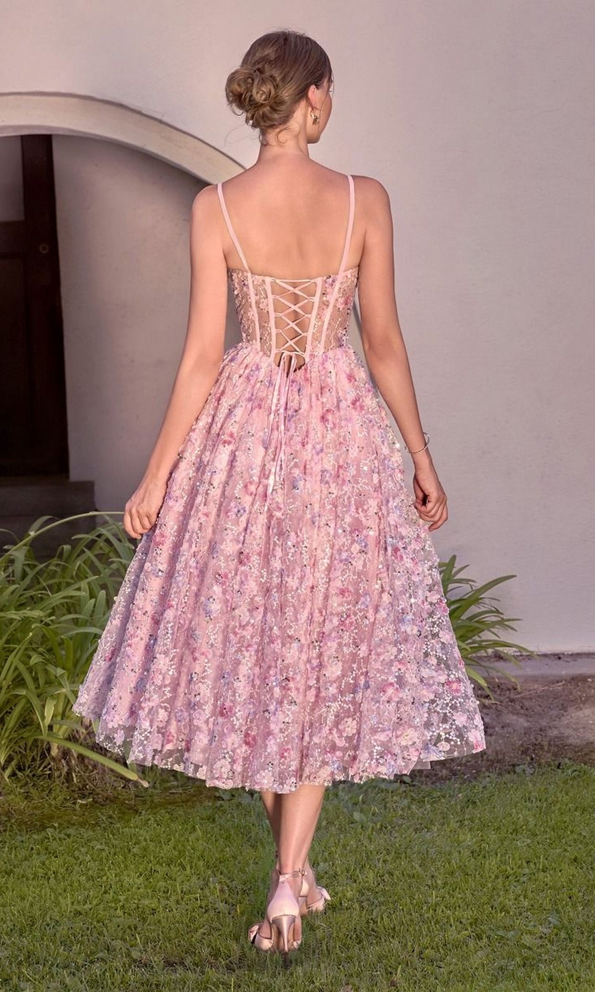 A woman wearing the Andrea & Leo A1523 midi prom dress with pink floral lace-up details, spaghetti straps, and a sheer corset bodice walks on grass toward a building with an arched doorway.