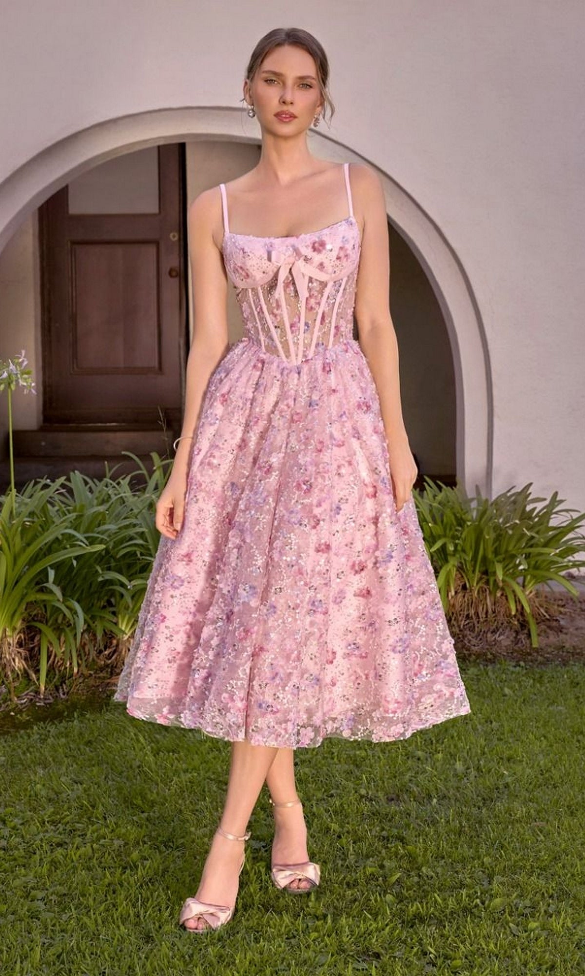 A woman stands on grass in front of an arched doorway, wearing the blush-colored “Midi Prom Dress: Andrea & Leo A1523,” which features a floral pattern, sheer corset bodice, and matching heels.