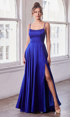 A woman stands indoors by large windows wearing the Lace-Up Classic Long Prom Dress B8402, a sleeveless royal blue A-line gown featuring a thigh-high slit.