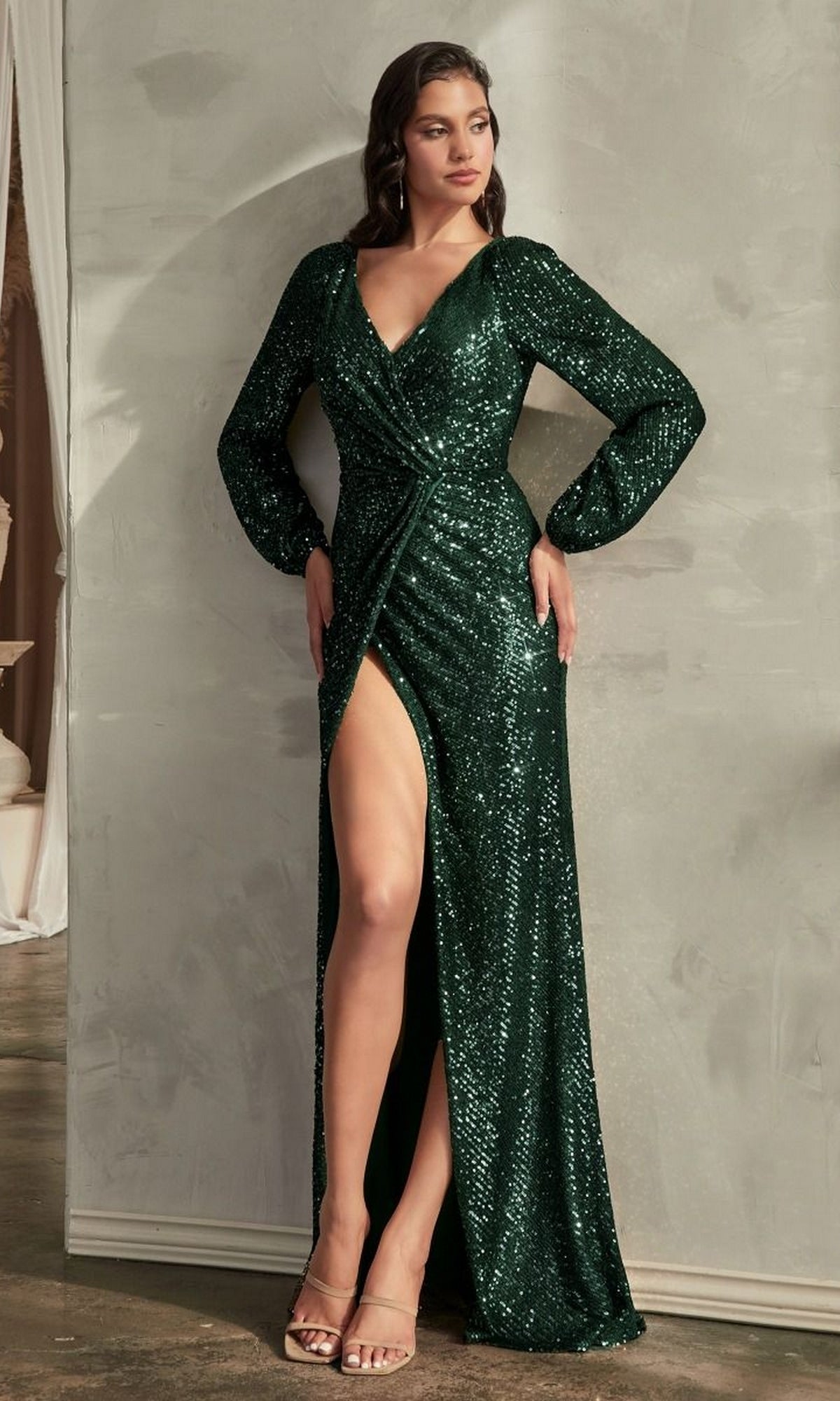 A woman stands indoors against a light wall wearing the Long Sleeve Long Sequin Formal Dress B8422—an elegant dark green gown featuring sequins, a V-neckline, and a high slit.