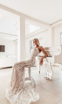Tina Holly wears the White Lace Bodycon Bridal Gown: BA999, sitting on a white bench beside a white grand piano in a bright, modern room with large windows.
