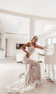 A woman in the White Lace Bodycon Bridal Gown: Tina Holly BA999 sits at a white grand piano in a bright, modern room with large windows.