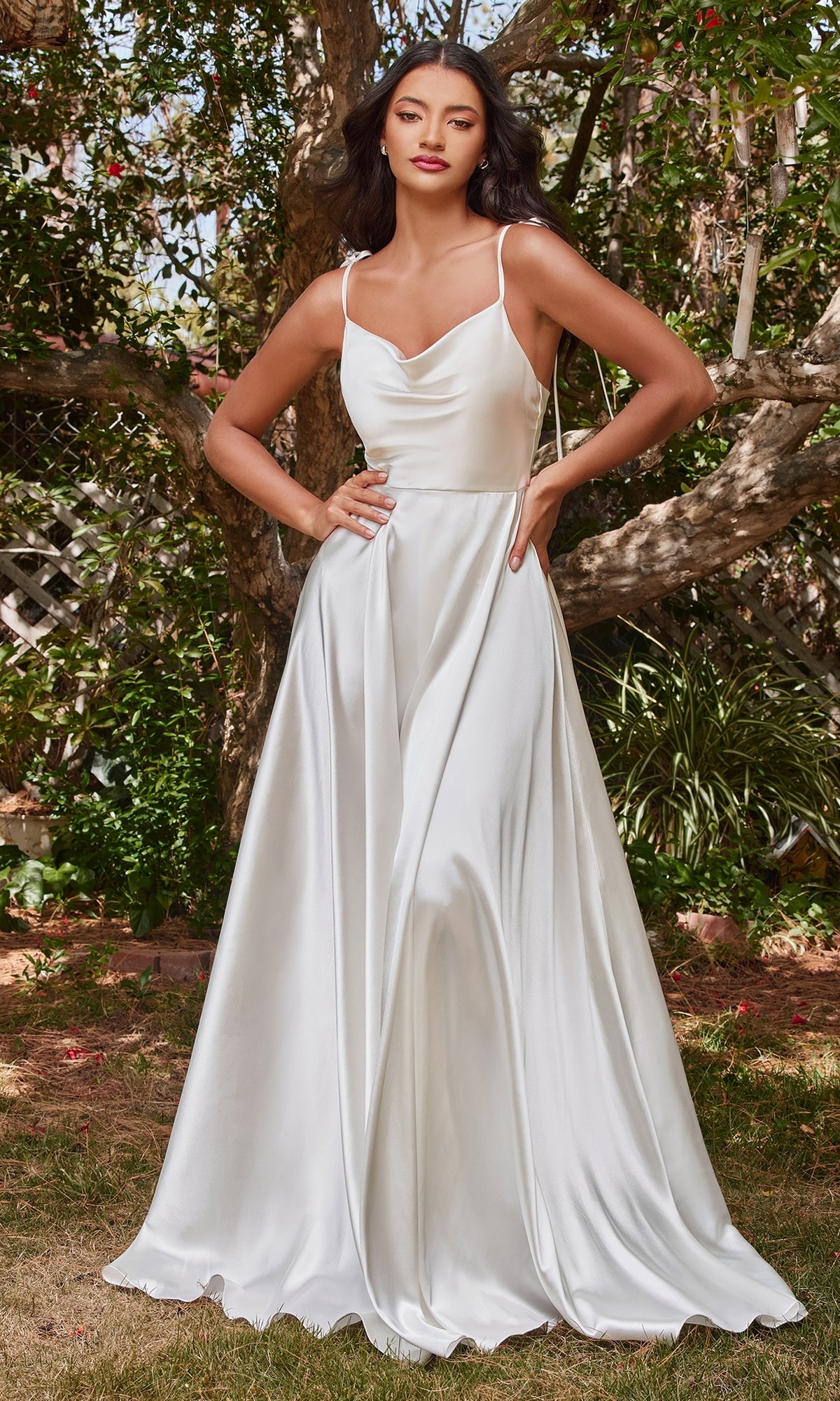 A woman stands outdoors surrounded by greenery, wearing the Long White Satin A-Line Formal Dress BD104W featuring thin straps and a cowl neckline.