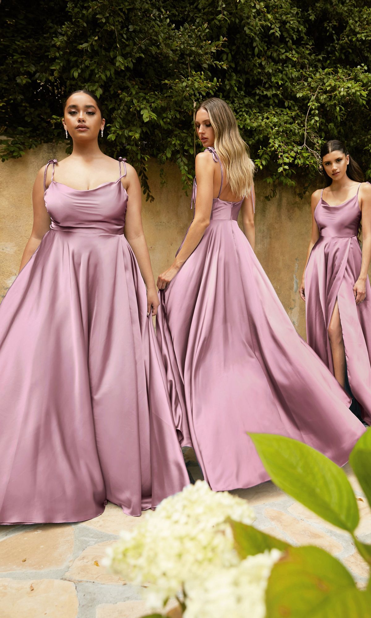 Three women wearing matching Ladivine BD104 cowl-neck long satin prom dresses stand outdoors on stone pavement, with greenery and a beige wall in the background.