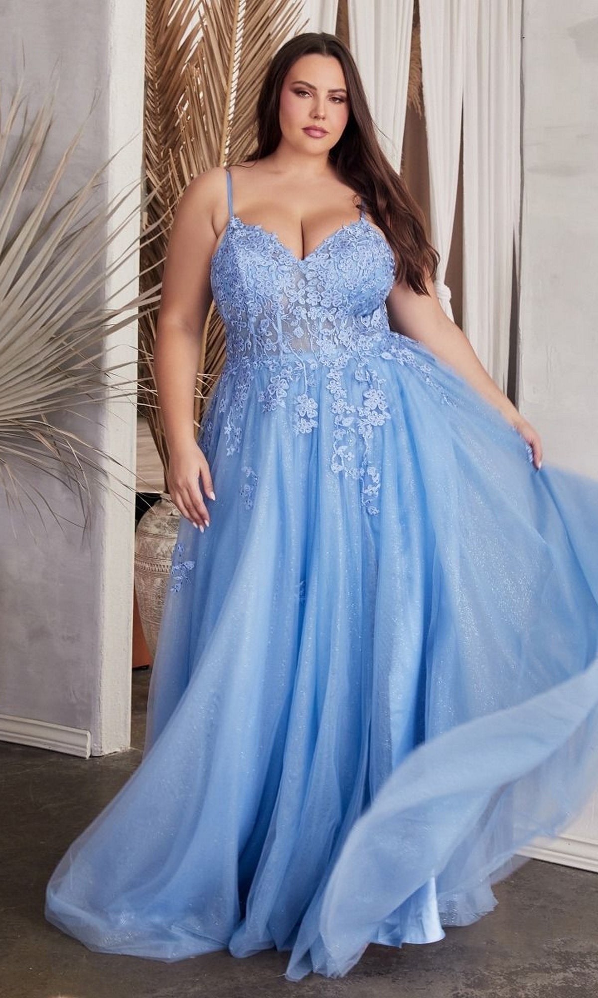 A woman in the Lace-Up Back Plus-Size Long Prom Ball Gown C148C, a light blue, floor-length embroidered lace dress with floral details, stands indoors and elegantly holds out part of the skirt.