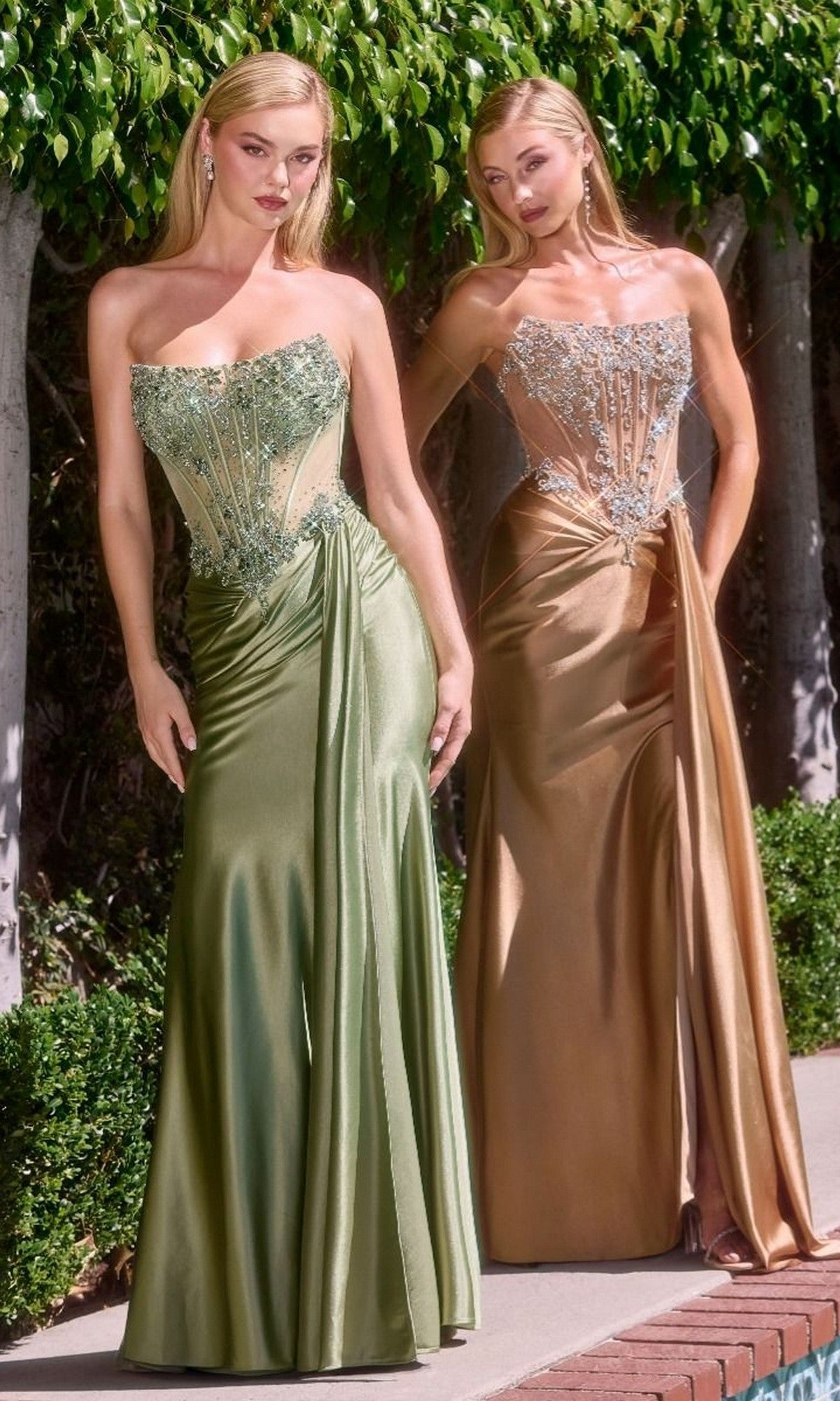 Two women stand outdoors among lush greenery, each wearing the Strapless Long Corset Prom Gown: Ladivine C169 with a fitted, beaded corset bodice—one in sage, the other in gold—posing gracefully amid trees.