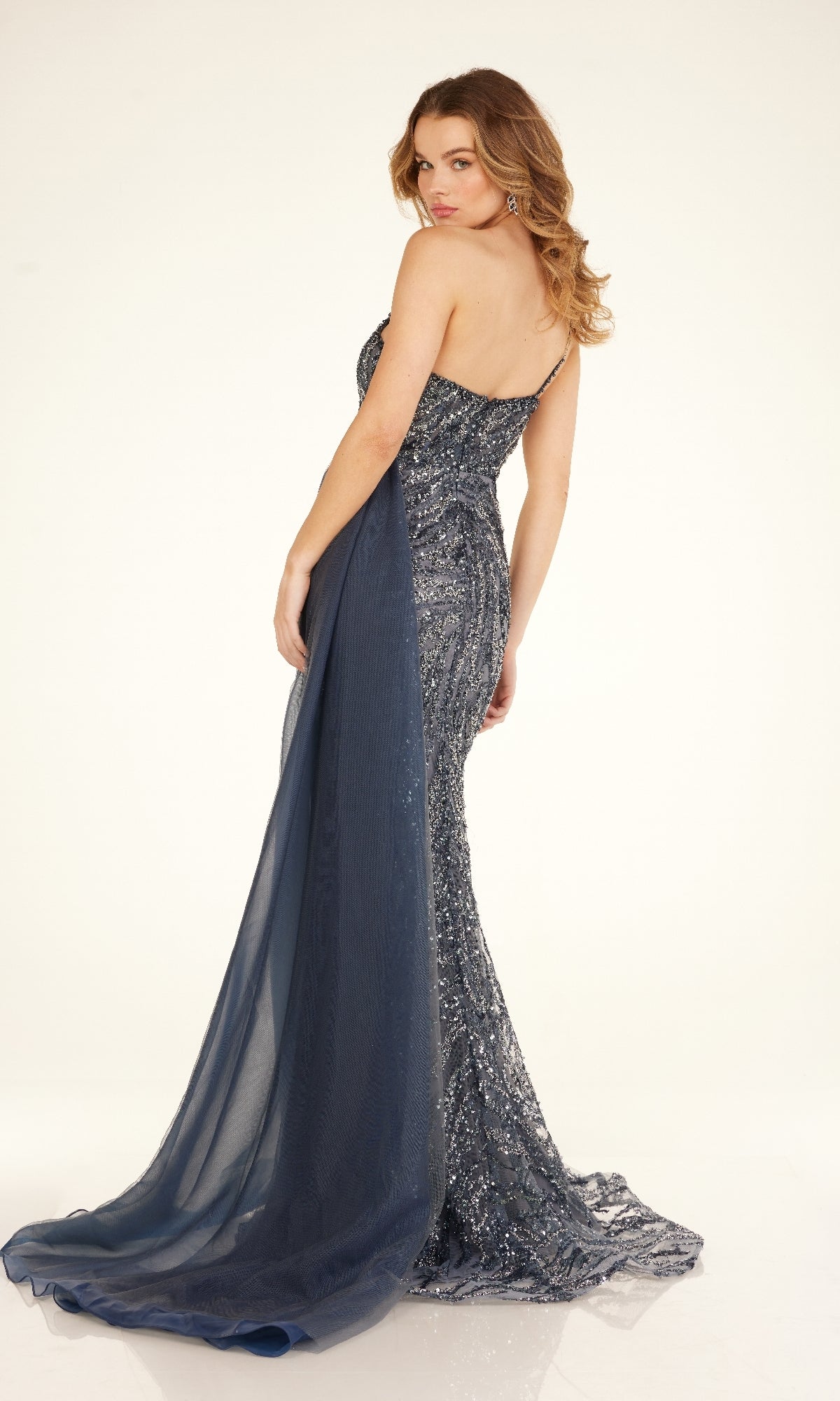A woman poses against a plain background wearing the Cecilia Couture C275, a navy blue, floor-length one-shoulder sequin prom dress with a sheer train.
