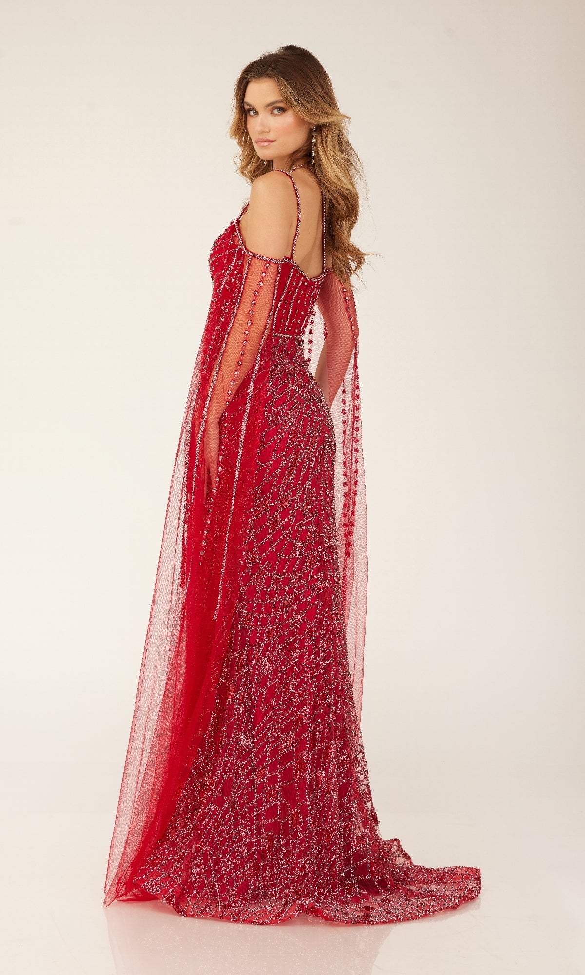 A woman poses in the Cecilia Couture C280, a fitted, floor-length sparkly formal dress with sheer draped details, embellishments, and a high leg slit, looking over her shoulder against a plain background.