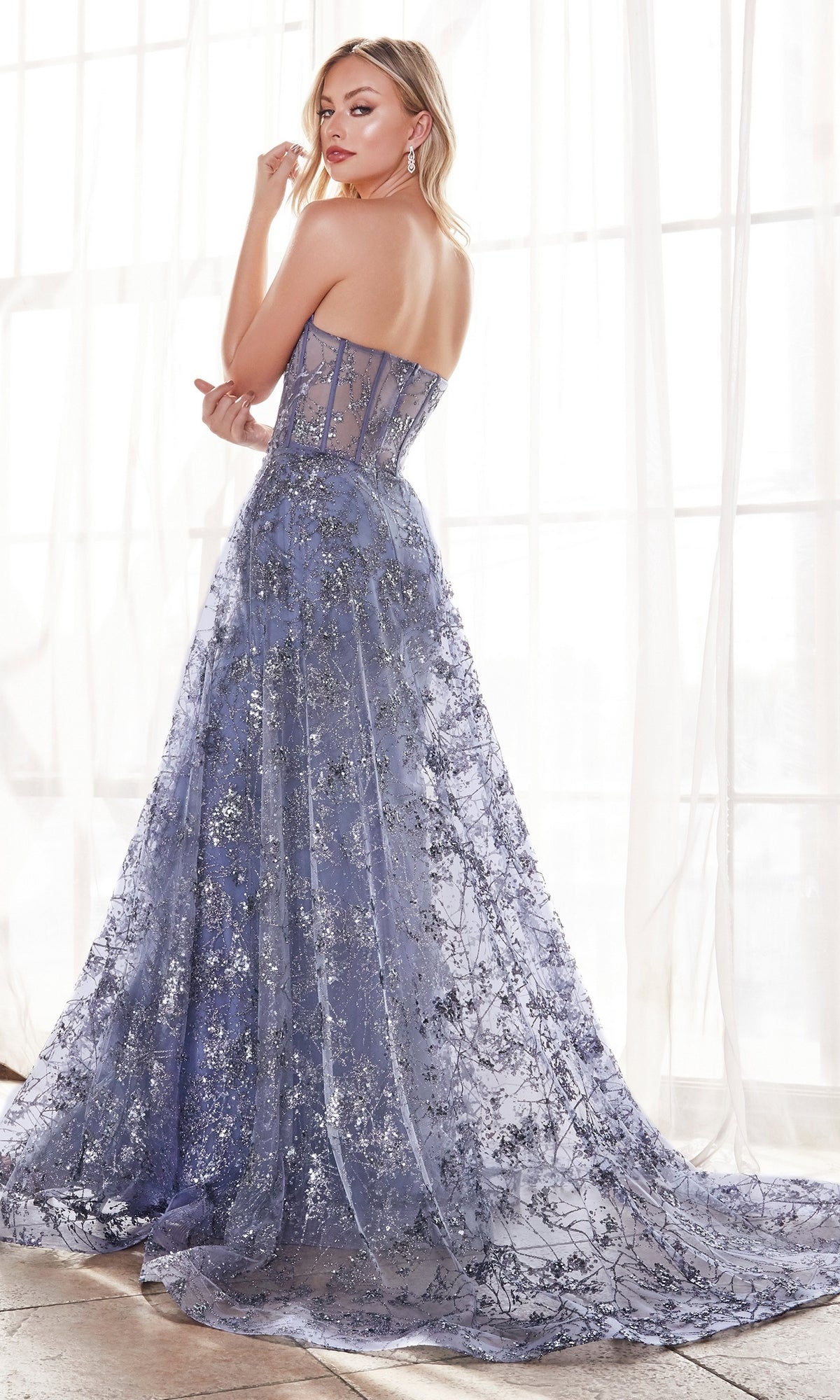 A woman stands before sheer curtains in the Glitter-Print Strapless Corset Prom Dress CB046, a lavender-blue gown with a sheer, embellished glitter-print bodice and a long flowing train.