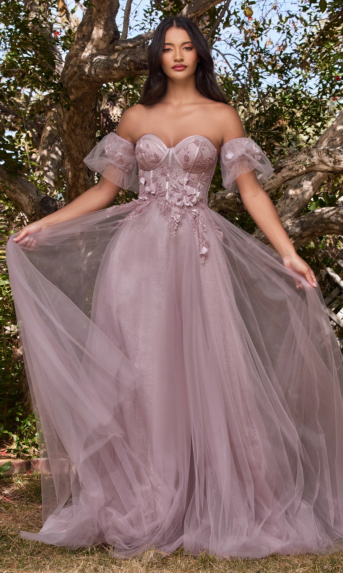 A woman stands outdoors wearing the Ladivine CB080 Glitter Tulle Ball Gown Prom Dress, featuring sheer tulle sleeves and a flowing, layered skirt.