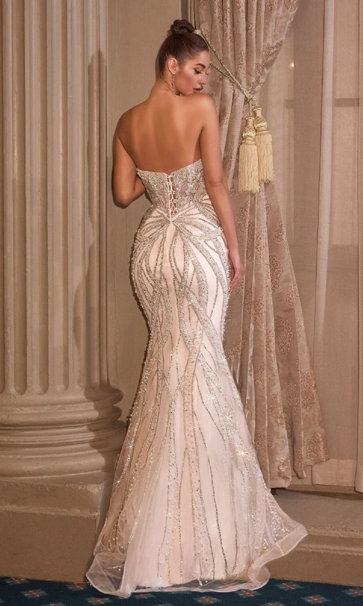 A woman stands with her back to the camera by a curtained window, showcasing the elegant silhouette of the Ladivine CC0688W long wedding dress adorned with silver crystal embellishments.