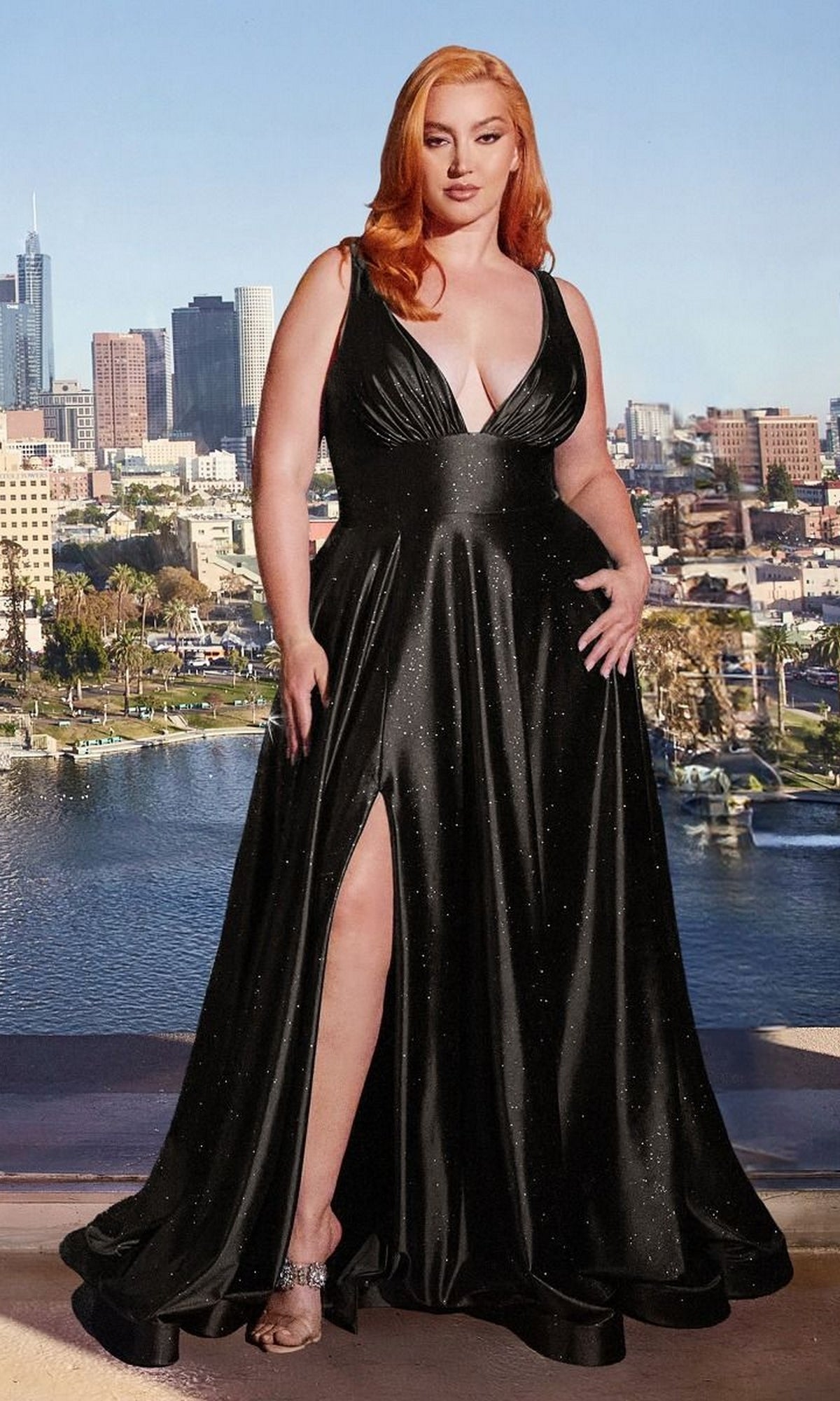 A woman with red hair in the Plus Long Glitter Prom Dress: Ladivine CC2349C, a black sleeveless gown with a thigh-high slit, stands before a city skyline and water.