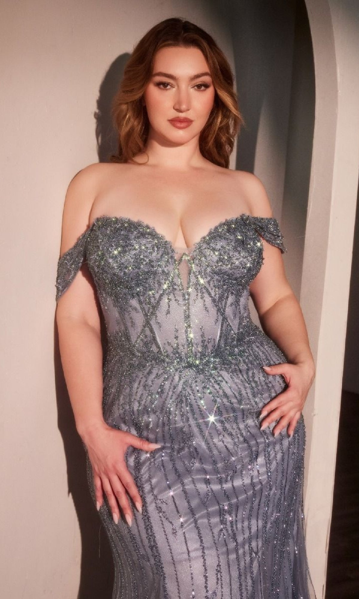 A woman poses with her hands on her hips against a light wall, wearing the Plus-size Blue Beaded Prom Gown: Ladivine CC341C.