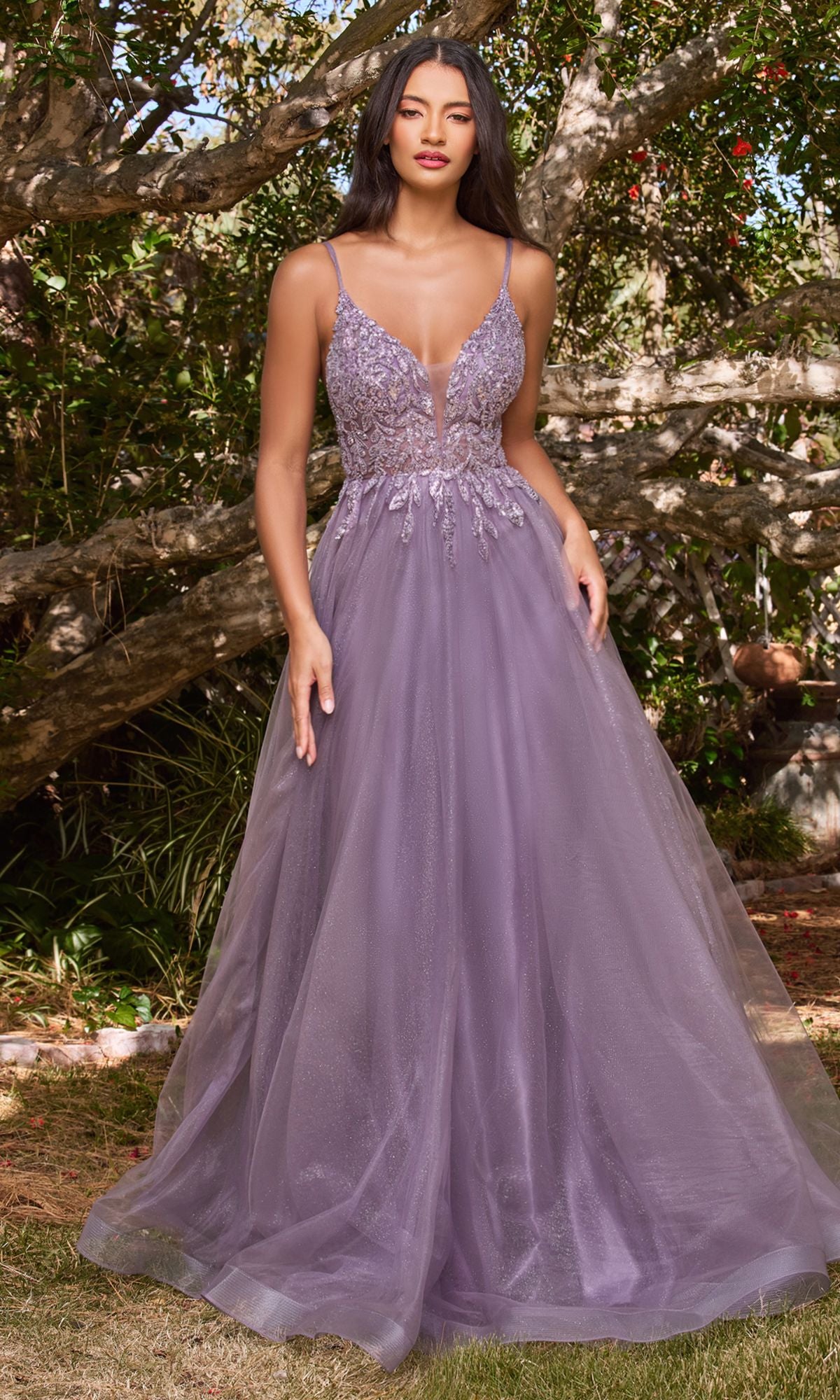 A woman stands outdoors in the Sheer-Corset Long Prom Ball Gown CD0154, a sleeveless, floor-length lavender dress with a beaded fitted bodice and flowing tulle skirt. Trees and greenery are visible in the background.