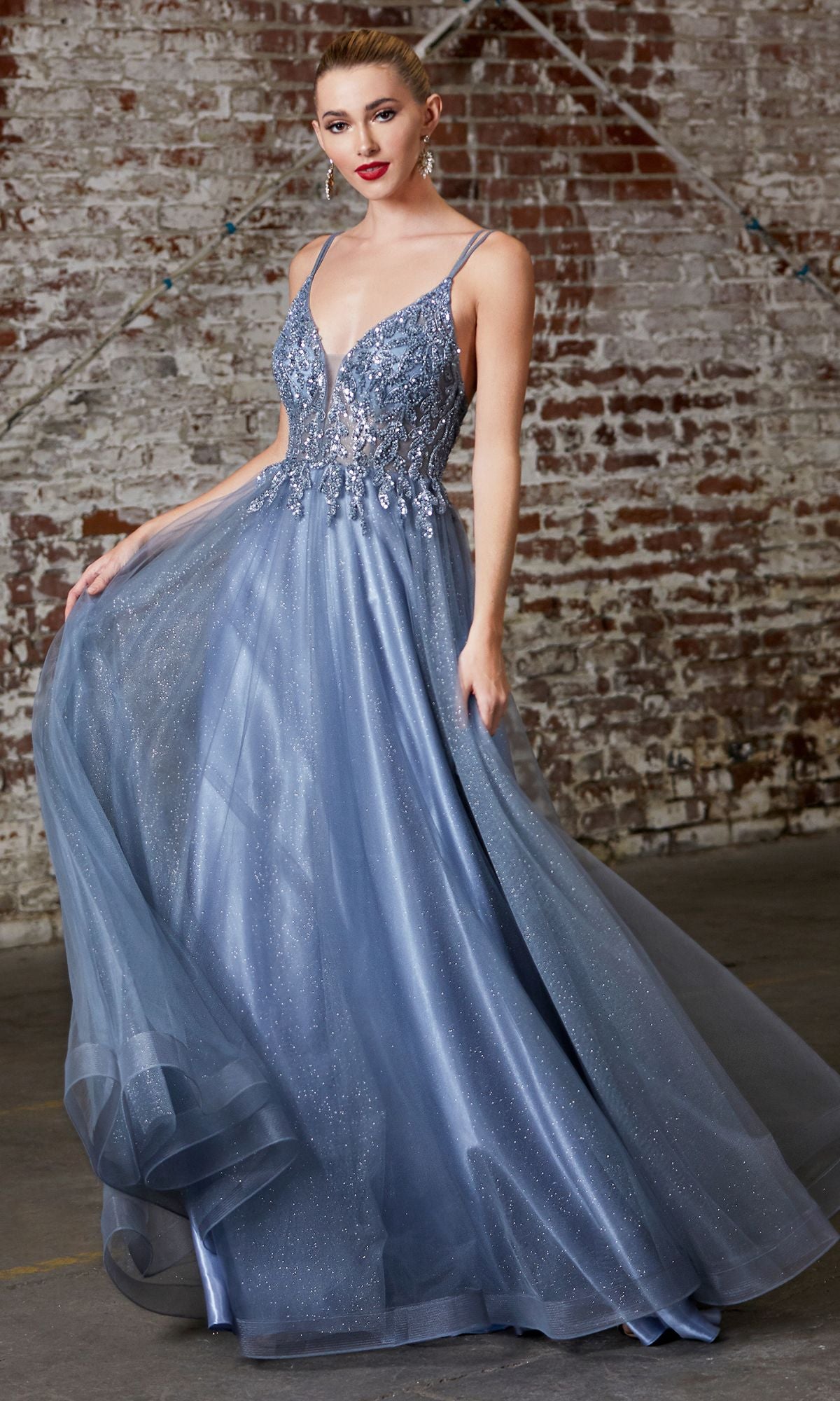 Woman wearing the Sheer-Corset Long Prom Ball Gown CD0154, featuring a sparkly blue fitted beaded bodice and flowing tulle skirt, stands in front of a weathered brick wall.
