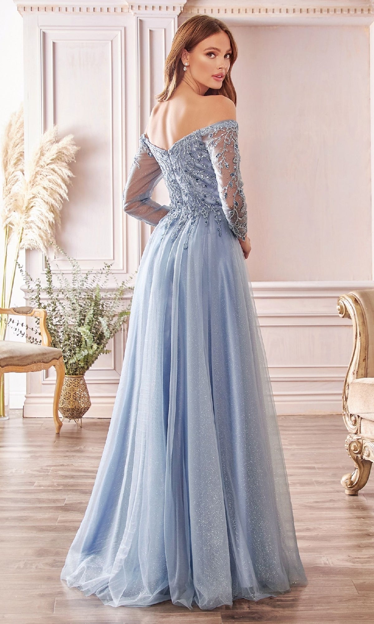 A woman stands indoors facing away, wearing the Ladivine CD0172 long sleeve prom dress in light blue with off-the-shoulder sheer sleeves and embroidered details.